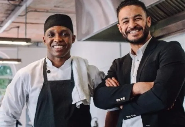 2 men in a kitchen one in chefs whites and one in a suit